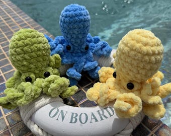 May include: Three crocheted octopus toys in shades of green, blue, and yellow, resting on a white life preserver. The life preserver has the words "ON BOARD" printed on it. The toys are placed near a pool.