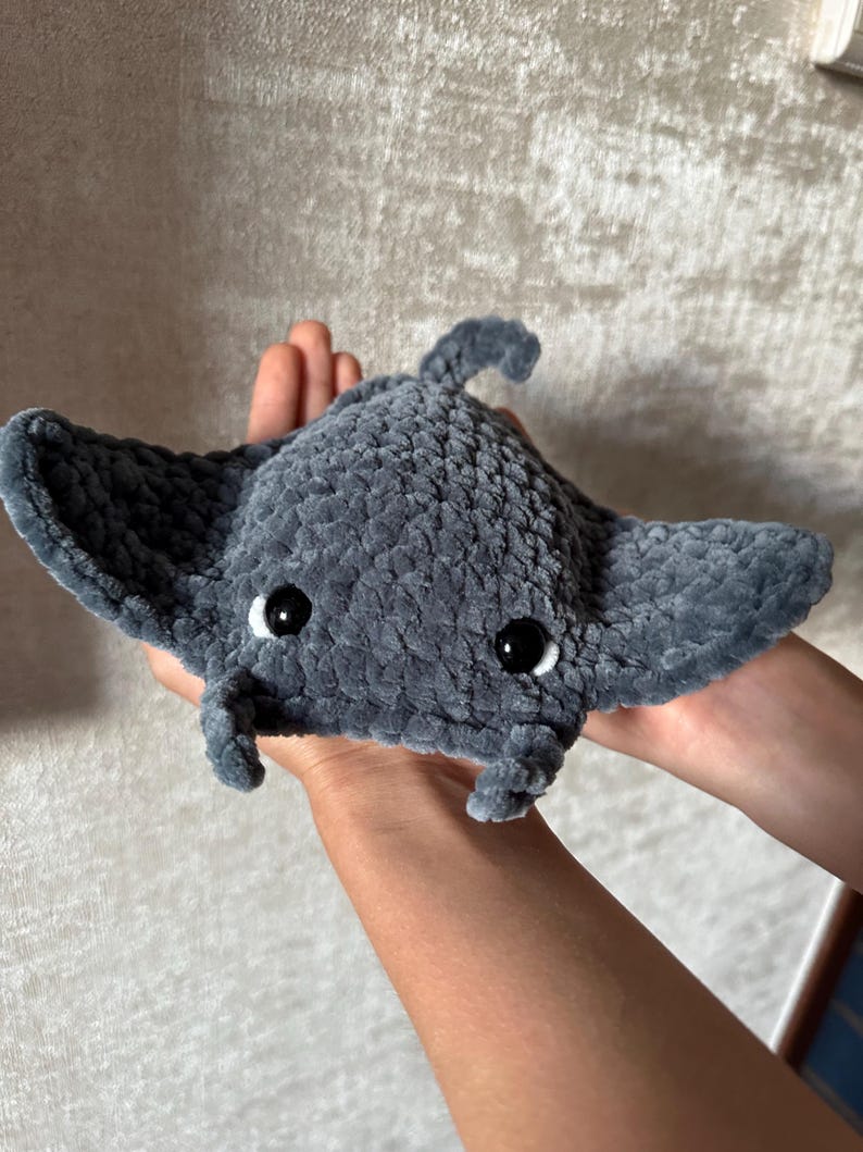 May include: A handmade, gray crocheted manta ray plush toy with black safety eyes. The plushie has a rounded body, wide wings, and a small tail. The toy is held in a person's hands.