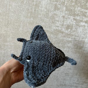 May include: A handmade, plush gray manta ray toy with black button eyes and small fins. The soft, textured fabric gives the toy a cuddly appearance. The manta ray is held in a hand against a neutral background.