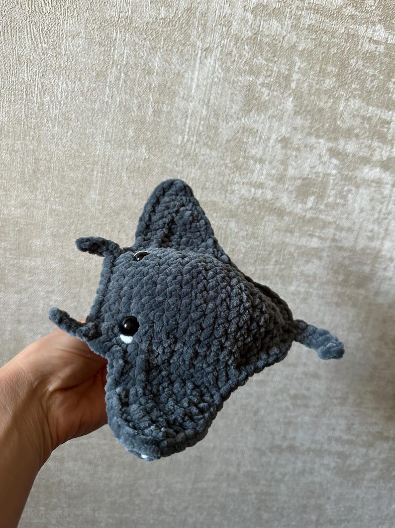 May include: A handmade, plush gray manta ray toy with black button eyes and small fins. The soft, textured fabric gives the toy a cuddly appearance. The manta ray is held in a hand against a neutral background.