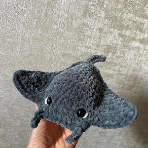 May include: A handmade, plush gray manta ray with black eyes and white accents. The soft, textured toy features a rounded body and outstretched wings, perfect for a child's room or ocean-themed decor.
