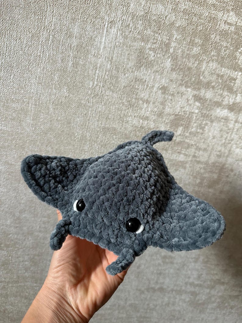 May include: A handmade, plush gray manta ray with black eyes and white accents. The soft, textured toy features a rounded body and outstretched wings, perfect for a child's room or ocean-themed decor.