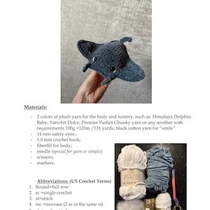 May include: A handmade amigurumi manta ray in shades of gray, with small black safety eyes. The plush toy is made with crochet techniques. The image also shows materials like yarn, a crochet hook, and scissors.