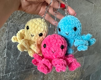 May include: Three crocheted octopus keychains in yellow, pink, and blue. Each plush octopus has black eyes and a smiling face. The keychains are attached to silver metal rings.