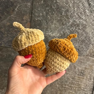 Puede incluir: Dos adornos de bellota tejidos a ganchillo con hilo suave y afelpado. Una bellota presenta una tapa de color beige claro con una base marrón dorada, mientras que la otra tiene una tapa marrón dorada con una base de color beige claro.