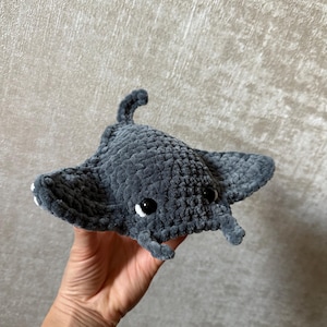 May include: A handmade gray crocheted manta ray plush toy with black safety eyes and small white accents. The plush has a textured surface and a small tail. The manta ray is a soft, cuddly toy.