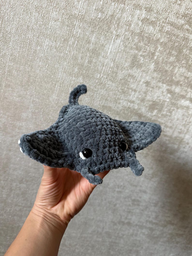 May include: A handmade gray crocheted manta ray plush toy with black safety eyes and small white accents. The plush has a textured surface and a small tail. The manta ray is a soft, cuddly toy.