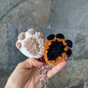 Pode incluir: Dois chaveiros de crochê em forma de pata, um marrom e branco, o outro laranja e preto, presos a um chaveiro prateado.