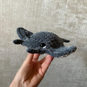 May include: A handmade, gray crocheted stingray plush toy with black safety eyes and white accents. The plush toy is held in a person's hand. The stingray has a soft, textured appearance.