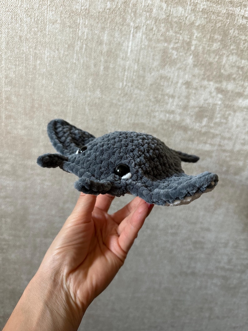 May include: A handmade, gray crocheted stingray plush toy with black safety eyes and white accents. The plush toy is held in a person's hand. The stingray has a soft, textured appearance.