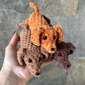Op de afbeelding: Drie handgemaakte gehaakte teckel honden in bruin- en oranjetinten. De honden hebben zwarte knoopogen en zijn gemaakt met een zacht, getextureerd garen. De teckels zijn een charmant, grillig knutselitem.