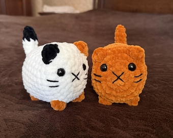 May include: Two handmade crochet cat figurines. One is white with black spots and orange accents, the other is orange. Both have black eyes and stitched facial features. The cats are small and spherical.