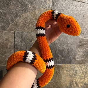 Puede incluir: Un juguete de peluche de serpiente naranja con rayas negras y blancas. La serpiente tiene una cabeza redondeada con pequeños ojos negros. El juguete está hecho de un material suave y texturizado.