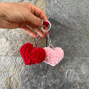 May include: Two heart-shaped keychains. One is a vibrant red, the other a soft pink. Both are made of a plush, textured material. Each keychain has a silver ring and chain.
