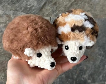 May include: Two small, round, crocheted hedgehog plush toys. One hedgehog has solid brown fuzzy yarn on its back, while the other features a mottled pattern of brown, white, and black fuzzy yarn. Both have white textured faces, black eyes, and black noses.