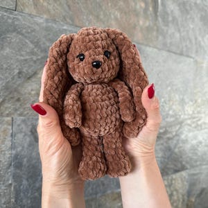 May include: A brown, plush bunny toy with long floppy ears, black eyes, and a small nose. The handmade stuffed animal is held in front of a neutral background. The bunny is made of a soft, textured material.