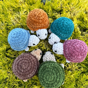 Puede incluir: Seis juguetes de tortuga de crochet en diferentes colores, incluyendo marrón, verde, azul, rosa y naranja. Las tortugas están dispuestas en círculo sobre un lecho de follaje verde.