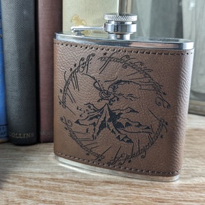 May include: A brown leather-bound flask with a silver top and a silver rim. The flask has an engraved design of a mountain range and the words "All that is gold does not glitter, not all those who wander are lost; the old that is strong does not wither, deep roots are not reached by the frost. From the ashes a fire shall be woken, a light from the shadows shall spring; renewed shall be blade that was broken, the crownless again shall be king."