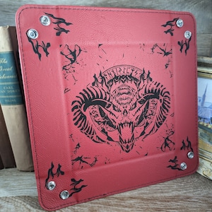 May include: Red leather dice tray with black stitching and a black and white graphic of a horned demon with the text "FINIS" and "LYX" on the top and bottom of the graphic. The tray has silver metal rivets around the edges.