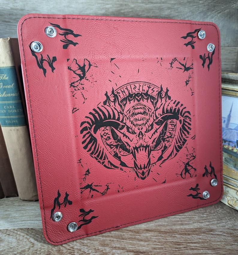May include: Red leather dice tray with black stitching and a black and white graphic of a horned demon with the text "FINIS" and "LYX" on the top and bottom of the graphic. The tray has silver metal rivets around the edges.