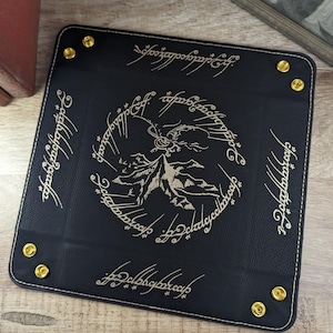 May include: Black leather dice tray with gold stitching and a gold embossed design of Mount Doom and the One Ring inscription in Elvish. The inscription reads "One Ring to rule them all, One Ring to find them, One Ring to bring them all and in the darkness bind them."