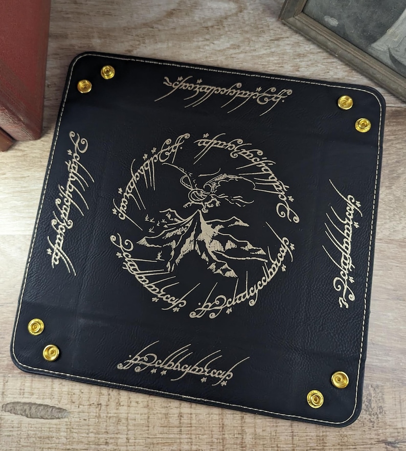 May include: Black leather dice tray with gold stitching and a gold embossed design of Mount Doom and the One Ring inscription in Elvish. The inscription reads "One Ring to rule them all, One Ring to find them, One Ring to bring them all and in the darkness bind them."