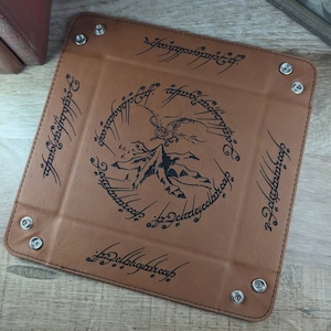 May include: A brown leather dice tray with a black inscription of the One Ring inscription from The Lord of the Rings. The inscription reads "One Ring to rule them all, One Ring to find them, One Ring to bring them all and in the darkness bind them." The tray has a black silhouette of Mount Doom and a dragon.
