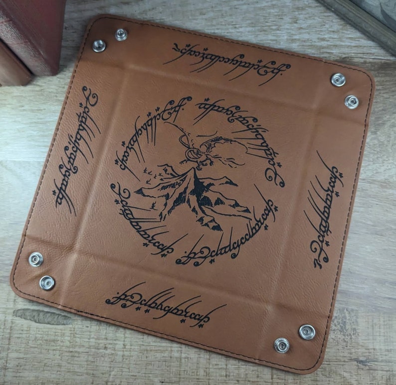 May include: A brown leather dice tray with a black inscription of the One Ring inscription from The Lord of the Rings. The inscription reads "One Ring to rule them all, One Ring to find them, One Ring to bring them all and in the darkness bind them." The tray has a black silhouette of Mount Doom and a dragon.