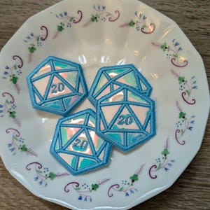 May include: Four blue and iridescent D20 dice patches with the number 20 embroidered on each. The patches are arranged on a white plate with a floral design.