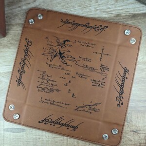 May include: A brown leather dice tray with a map of Middle-earth from The Lord of the Rings printed on it. The tray has a stitched border and four metal rivets. The text "One Ring to rule them all, One Ring to find them, One Ring to bring them all and in the darkness bind them" is printed around the edge of the tray.