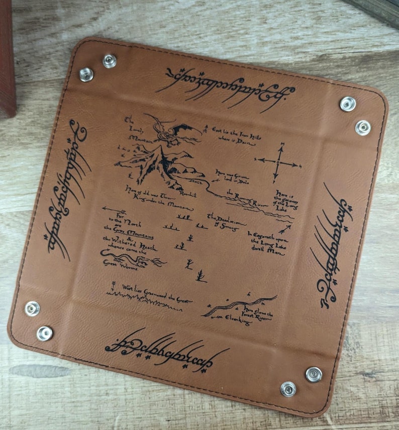 May include: A brown leather dice tray with a map of Middle-earth from The Lord of the Rings printed on it. The tray has a stitched border and four metal rivets. The text "One Ring to rule them all, One Ring to find them, One Ring to bring them all and in the darkness bind them" is printed around the edge of the tray.
