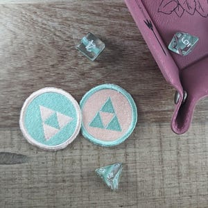 May include: Two embroidered patches with a triangular design in shades of teal and pink. The patches are on a wooden surface with two dice.