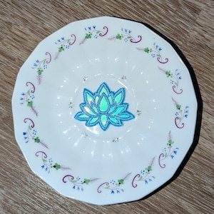 May include: White ceramic plate with a scalloped edge, featuring a central iridescent lotus flower design. The rim is decorated with green, blue, and purple floral patterns. The plate has a diameter of approximately 8 inches.