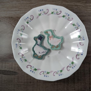 May include: Two embroidered goose patches rest on a white, scalloped-edge plate with a floral border. One goose wears a top hat, the other a pink bow. The plate sits on a wooden surface.