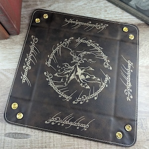 May include: A brown leather dice tray with gold accents and the inscription "One Ring to rule them all, One Ring to find them, One Ring to bring them all and in the darkness bind them" in Elvish script. The tray features a mountain and a dragon.