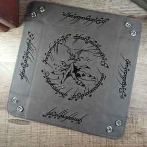 May include: A gray leather tray with a black inscription of the One Ring inscription from The Lord of the Rings. The inscription reads "One Ring to rule them all, One Ring to find them, One Ring to bring them all and in the darkness bind them." The tray also features a black silhouette of Mount Doom and a dragon.
