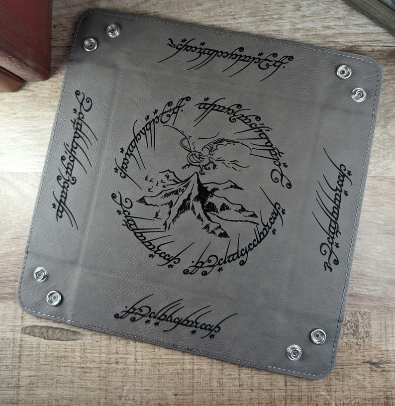 May include: A gray leather tray with a black inscription of the One Ring inscription from The Lord of the Rings. The inscription reads "One Ring to rule them all, One Ring to find them, One Ring to bring them all and in the darkness bind them." The tray also features a black silhouette of Mount Doom and a dragon.