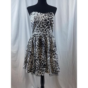 Puede incluir: Vestido sin tirantes con estampado de animales en blanco y negro. El vestido presenta un corpiño ajustado y una falda acampanada. La falda tiene una capa de tul negro en el dobladillo. El vestido se exhibe en un maniquí negro.