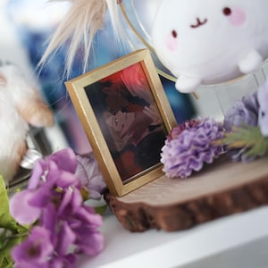 Puede incluir: Una composición decorativa con una pequeña obra de arte enmarcada con un marco dorado, un peluche y flores moradas. La obra de arte está sobre una rodaja de madera, sobre un fondo blanco. El peluche es blanco con una cara de dibujos animados.