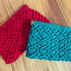 May include: Two macrame clutches in red and teal. The clutches have a woven pattern and a rectangular shape. The red clutch is on the left, and the teal clutch is on the right. Both clutches have a metal clasp.