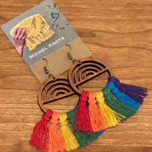 Könnte beinhalten: Holz-Creolen mit Regenbogen-Design und bunten Quasten. Die Ohrringe haben ein lasergeschnittenes Regenbogenmuster und sind an bronzefarbenen Ohrhaken befestigt. Die Quasten sind in den Farbtönen Rot, Orange, Gelb, Grün, Blau und Lila gehalten.