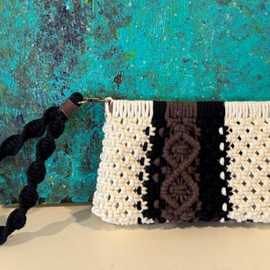 Pode incluir: Uma bolsa de mão de macramé branca e preta com um design tecido castanho. A bolsa tem uma alça trançada preta.