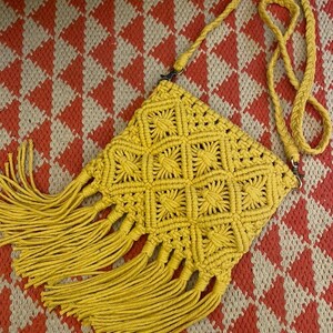 May include: A yellow macrame crossbody bag with a geometric pattern and fringe detail. The bag has a long, braided strap and is set against a red and cream diamond pattern.