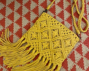 May include: A yellow macrame crossbody bag with a geometric pattern and fringe detail. The bag has a long, braided strap and is set against a red and cream diamond pattern.