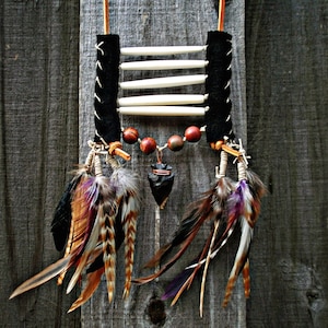 May include: A Native American-inspired necklace with a black leather strap, bone beads, and feathers. The necklace features a dark stone pendant and red and brown beads.
