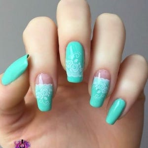 May include: Close-up of a hand with five fingernails painted in a light turquoise colour. Each nail has a white lace design with floral accents.