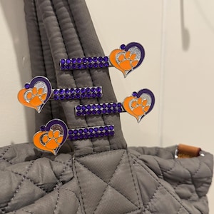 Peut inclure: Barrettes en forme de cœur avec un motif de patte orange et des accents violets. Les barrettes sont fixées à un sac fourre-tout matelassé gris. Les barrettes sont ornées de strass violets.