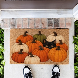 May include: A rectangular coir doormat featuring a printed illustration of various pumpkins in shades of orange, green, and cream, with a black cat with bright yellow eyes peeking out from behind the pumpkins in the center of the mat.