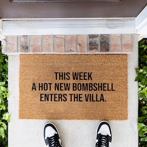 May include: A rectangular tan coir doormat featuring black text that reads: THIS WEEK A HOT NEW BOMBSHELL ENTERS THE VILLA. The mat is made of natural coconut fiber and is positioned on a concrete entryway floor in front of a dark brown door.