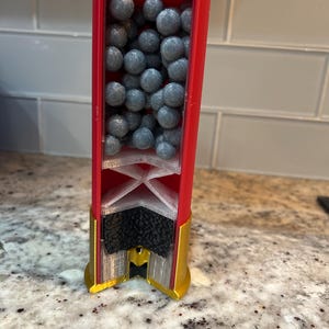May include: A cutaway view of a red and gold shotgun cartridge, showing the internal structure. The cartridge is packed with numerous grey pellets. The base of the cartridge is gold, and the internal parts are white and black.